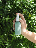A hand holding a 50ml Fragrant Room Spray bottle with a green label, in front of a background of green leaves.