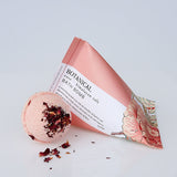 A pink botanical bath bomb with dried rose petals, next to its packaging tube which is also shown. The packaging has floral designs and the product is labelled 'BOTANICAL BATH BOMB'.
