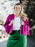 A woman smiling and wearing a pink cashmere blend coat with kimono style sleeves over a white top and a green skirt.